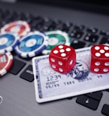 A set of red dice and poker chips are placed on top of a credit card, which is situated on a laptop keyboard. The poker chips are in various colors, including blue, green, red, and white.