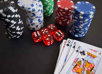 Stacks of poker chips in various colors including black, white, red, blue, and green are accompanied by red dice and a hand of playing cards featuring face cards such as the Queen and King of Clubs.