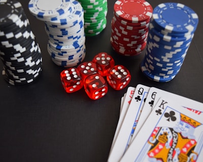 Stacks of poker chips in various colors including black, white, red, blue, and green are accompanied by red dice and a hand of playing cards featuring face cards such as the Queen and King of Clubs.