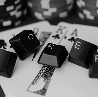 A series of playing cards and poker chips are displayed. The cards, including a ten, jack, queen, king, and ace of clubs, are spread out with black keyboard keys arranged to spell the word 'POKER'. The image is in black and white, giving it a classic, stylish look.
