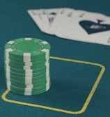 A stack of green poker chips is positioned on a green felt table within a yellow rectangular outline. In the background, there is a fanned set of playing cards, including an ace of spades visible.