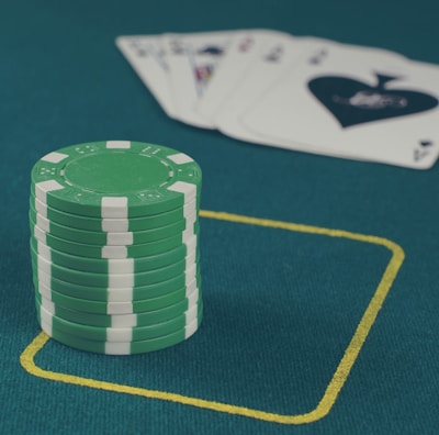 A stack of green poker chips is positioned on a green felt table within a yellow rectangular outline. In the background, there is a fanned set of playing cards, including an ace of spades visible.