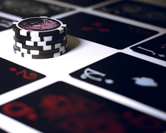 A stack of poker chips with a distinctive black and white pattern is placed on a table covered with several playing cards. The cards are dark with red and white symbols, creating a bold contrast against the lighter surface.