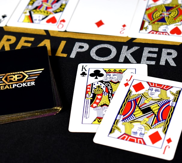 Poker-themed setup featuring a deck of cards on a table with the words 'REAL POKER' visible. Two cards, the King of Clubs and Jack of Diamonds, are prominently displayed. The design is vibrant, with detailed illustrations on the cards.