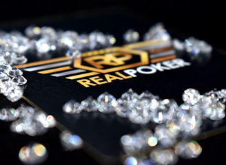 A collection of sparkling, clear gemstones surrounds a dark surface featuring the prominent logo and text 'Real Poker.' The logo design includes gold and white elements set against a black background.
