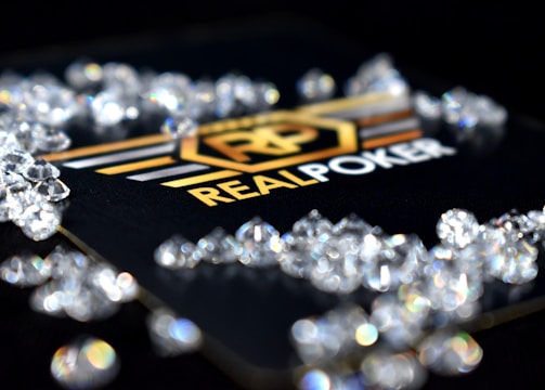 A collection of sparkling, clear gemstones surrounds a dark surface featuring the prominent logo and text 'Real Poker.' The logo design includes gold and white elements set against a black background.
