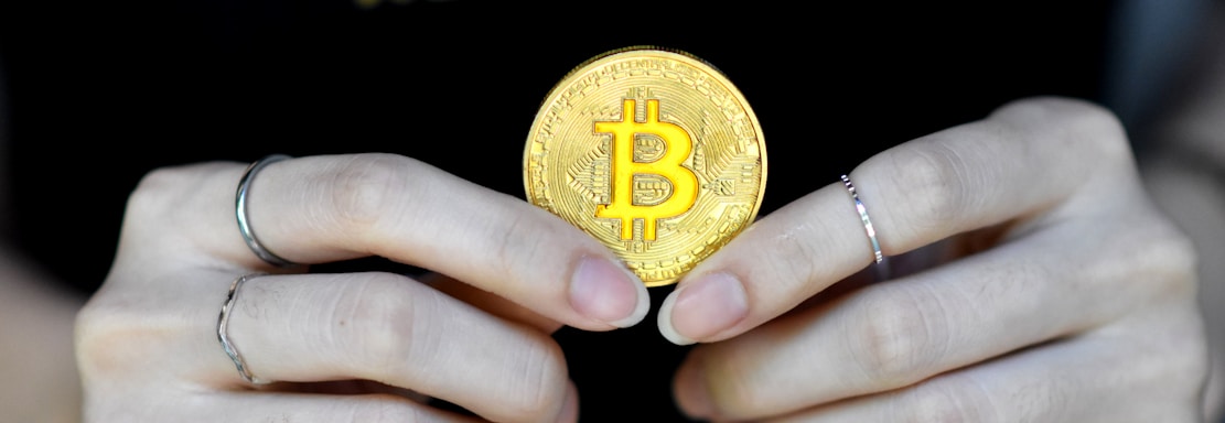A person holds a golden coin with a Bitcoin symbol between their fingers. The background features the word 'REALPOKER' in bold letters on a dark shirt.