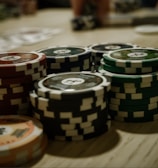Stacks of poker chips are arranged on a wooden table, with various denominations and colors such as red, green, and black. Some playing cards are blurred in the background, indicating a game setting.