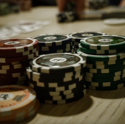 Stacks of poker chips are arranged on a wooden table, with various denominations and colors such as red, green, and black. Some playing cards are blurred in the background, indicating a game setting.
