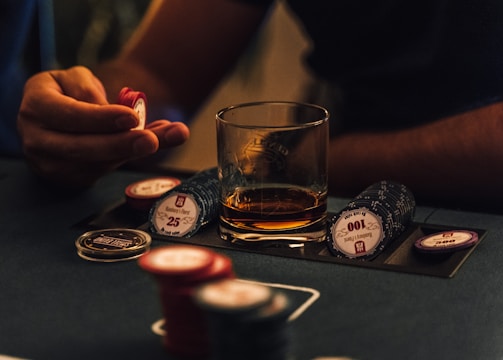 A hand holding several poker chips, with a glass of whiskey and a stack of poker chips on a table. The setting appears to be a dimly lit environment, suggesting an intimate or focused atmosphere.