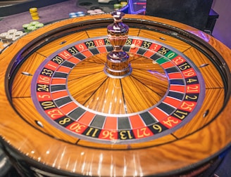 A roulette wheel with alternating red and black numbered pockets on a wooden platform. The wheel contains a small white ball resting in one of the pockets, indicating gameplay. Surrounding the wheel are stacks of colorful poker chips, suggesting a casino environment.