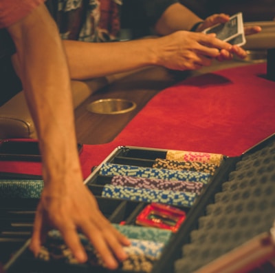 A person reaches into a poker chip case on a red felt table, while another person holds playing cards. The scene is dimly lit, creating a warm and engaged atmosphere.