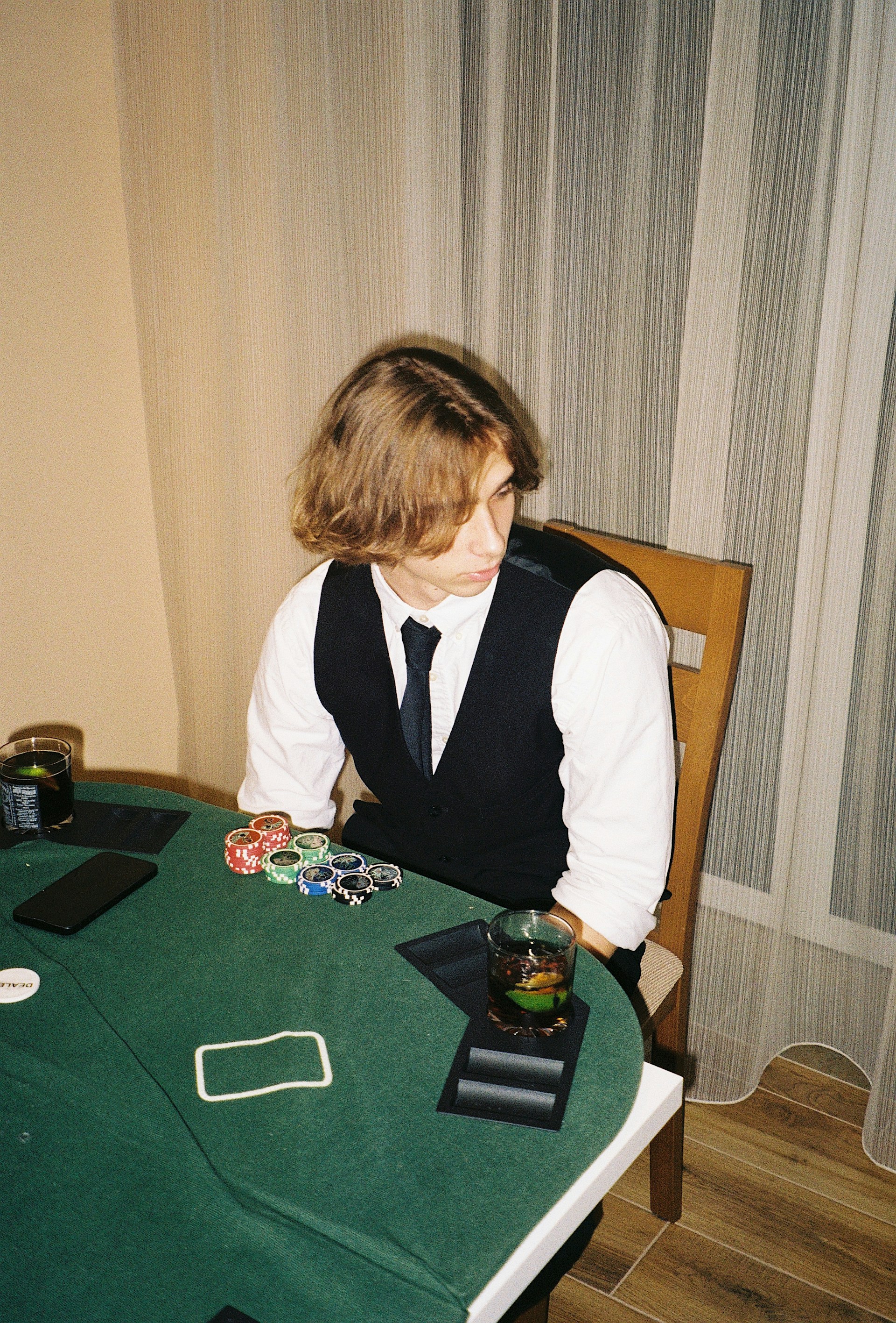 A person with shoulder-length hair is sitting at a poker table. They are wearing a white shirt, black vest, and tie, with an array of poker chips in front of them. There are two glasses with drinks on the table, and a smartphone is placed nearby. The background consists of vertical striped curtains.