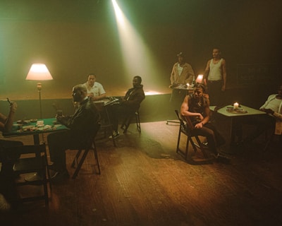 Several people sit at tables in a dimly lit room with a warm, moody atmosphere. A lamp and candle provide soft lighting, casting shadows across their faces. Cards and chips are on the tables, suggesting a game of poker. The setting is rustic, with wooden floors and simple furniture.
